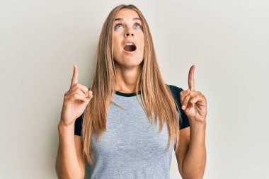 Gündelik kıyafetler giyen genç sarışın kadın şaşırmış ve şaşırmış bir şekilde yukarı bakıyor ve parmaklarıyla işaret ediyor ve kollarını kaldırıyor.. 