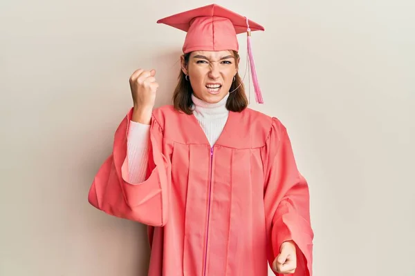 卒業キャップと儀式用のローブを身に着けている若い白人女性は怒りと怒りで叫んでいる間に怒りと怒りに満ちた拳を上げ怒っています 怒りと攻撃的な考え方 — ストック写真