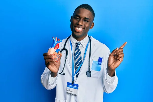 Молодой Африканский Американец Форме Доктора Держит Сердце Улыбаясь Счастливо Указывая — стоковое фото