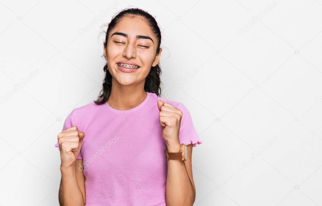 Hispanic teenager girl with dental braces wearing casual clothes celebrating surprised and amazed for success with arms raised and eyes closed. winner concept. 