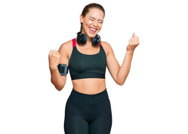 Güzel sarışın kadın spor kıyafetleri giyiyor ve kulaklık takıyor. Çok mutlu ve heyecanlı. Kazanan jestler yapıyor, kollarını kaldırıyor, gülümsüyor ve başarı için bağırıyor. kutlama konsepti. 