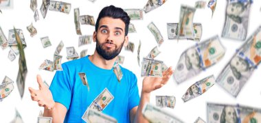 Sakallı, sıradan tişörtsüz, kafası karışmış, kolları ve elleri havada genç ve yakışıklı bir adam. şüphe kavramı.