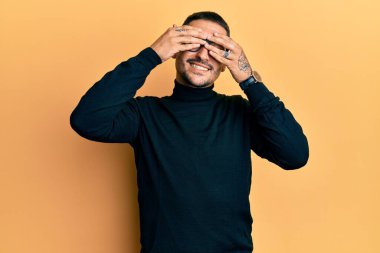 Balıkçı yaka kazak ve gözlük takan, gözleri kapayan neşeli ve komik gülümseyen yakışıklı bir adam. kör kavram. 