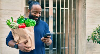 Sakallı Afrikalı Amerikalı adam elinde süpermarketten alınmış kağıt torbalarla akıllı telefon kullanıyor.