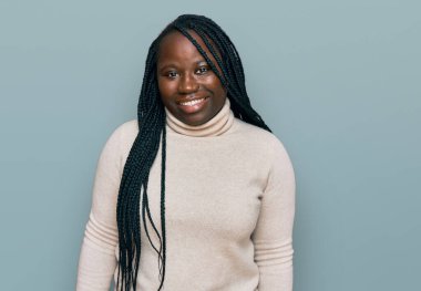 Saçları örgülü genç siyah bir kadın günlük kışlık süveter giyiyor. Olumlu ve mutlu görünüyor. Kendine güvenen bir gülümsemeyle dişlerini gösteriyor. 