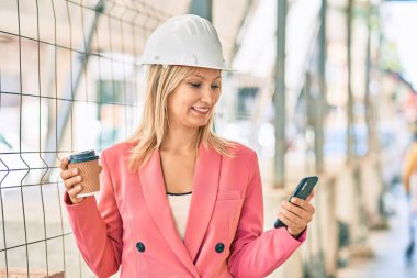 Genç beyaz mimar kadın akıllı telefon kullanıyor ve şehirde kahve içiyor.