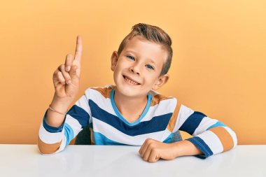 Beyaz elbiseli sevimli bir çocuk masanın üzerine oturmuş kendini beğenmiş ve mutlu bir şekilde gülümserken bir numaralı parmağıyla işaret ediyor.. 