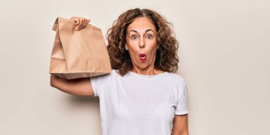 Orta yaşlı bir kadın elinde paketle beyaz arka planda yemek dağıtıyor. Şaşırmak için ağzı açık kalmış, korkmuş ve şaşırmış. İnanmayan yüz.