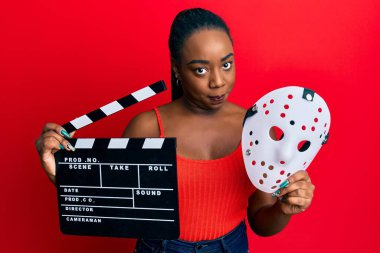 Genç Afro-Amerikan kadın elinde video film panosu ve korku maskesi tutuyor şüpheci ve sinirli, sorun yüzünden surat asıyor. negatif kişi. 