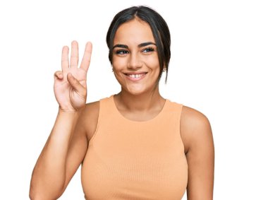 Genç esmer kadın gündelik giysiler giyip üç numaralı parmaklarıyla işaret ederken kendinden emin ve mutlu bir şekilde gülümsüyor.. 