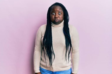 Saçları örgülü, kışlık süveter giyen, komik yüzlü, yanak şişiren genç siyahi bir kadın. Havayla şişmiş ağız, çılgın bir ifade.. 