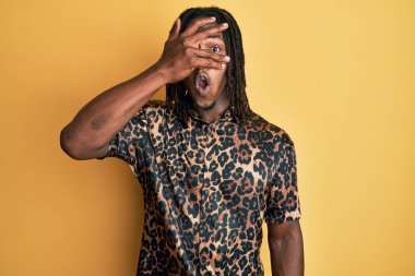 Saçları örgülü, leopar desenli hayvan desenli tişörtlü Afro-Amerikan bir adam. Şok içinde, yüzünü ve gözlerini kapayarak utangaç bir ifadeyle parmaklarından bakıyor.. 