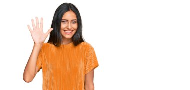 Genç, esmer, Arap bir kadın, günlük kıyafetler giyiyor. Beş numarayı gösteriyor ve parmaklarıyla işaret ediyor. Kendinden emin ve mutlu bir şekilde gülümsüyor.. 