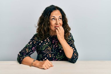 Masada rahat kıyafetler giyen güzel orta yaşlı bir kadın gergin ve gergin görünüyor. Ağzı tırnak yiyen ellerle. anksiyete. 