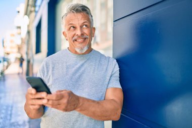 Orta yaşlı, ispanyol, kır saçlı, mutlu gülümseyen adam şehirde akıllı telefon kullanıyor..