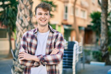 Kollarını kavuşturmuş genç beyaz adam şehirde mutlu bir şekilde gülümsüyor..