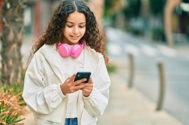 Güzel İspanyol kız şehirde akıllı telefon ve kulaklık kullanarak mutlu bir şekilde gülümsüyor..