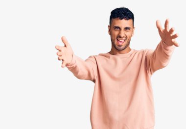 Gündelik giysiler giymiş genç İspanyol adam kameraya bakıp sarılmak için kollarını açmış gülümsüyor. Mutluluğu kucaklayan neşeli bir ifade.. 