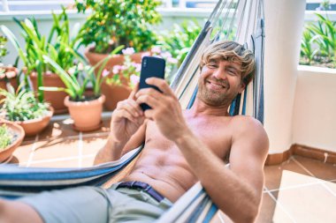 Orta yaşlı yakışıklı bir adam evinin terasında, bir hamakta telefonla dinleniyor.