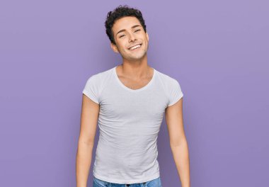 Genç ve yakışıklı bir adam günlük beyaz tişört giyiyor. Pozitif ve mutlu görünüyor. Kendine güvenen bir gülümsemeyle dişlerini gösteriyor. 