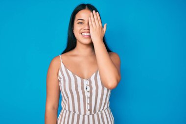 Genç, güzel Asyalı bir kız günlük kıyafetler giyiyor. Bir gözünü eliyle kapatıyor. Yüzünde kendine güvenen bir gülümseme ve sürpriz bir duygu var.. 