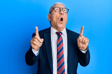 İş elbisesi giymiş, kravatını takmış beyaz bir adam şaşırmış ve şaşırmış bir şekilde yukarı bakıyor ve parmaklarıyla işaret ediyor ve kollarını kaldırıyor.. 