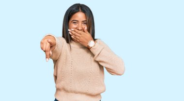 Kışın yün kazak giyen genç Latin kız sana gülüyor, eli ağzında kamerayı işaret ediyor, utanç ifadesi var. 