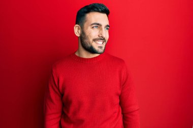 Latin kökenli genç bir adam günlük giysiler giyiyor, rahat bir görünüm takınıyor, doğal bir yüz ifadesi ve kendinden emin bir gülümsemeyle poz veriyor.. 