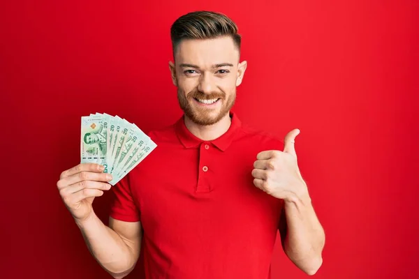 Joven Pelirrojo Sosteniendo Billetes Tailandeses Baht Sonriendo Feliz Positivo Pulgar —  Fotos de Stock