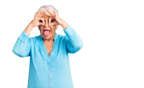 年长的漂亮女人 蓝眼睛 灰头发 穿着夏装 像双筒望远镜一样做手势 伸出舌头 用手指看东西 疯狂的表达 — 图库照片