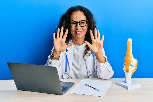 Beautiful middle age woman doctor at orthopedic clinic showing and pointing up with fingers number ten while smiling confident and happy.