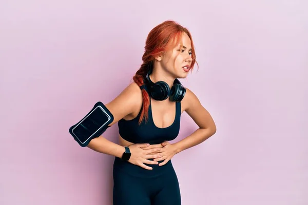 Young beautiful redhead woman wearing gym clothes and using headphones with hand on stomach because nausea, painful disease feeling unwell. ache concept.