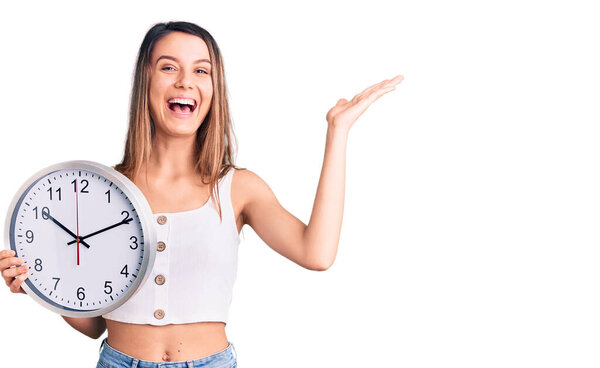 Молодая красивая девушка с большими часами празднования победы с счастливой улыбкой и выражение победителя с поднятыми руками 