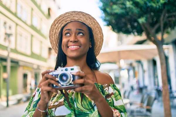 Young african american tourist woman on vacation smiling happy using vintage camera at the city.