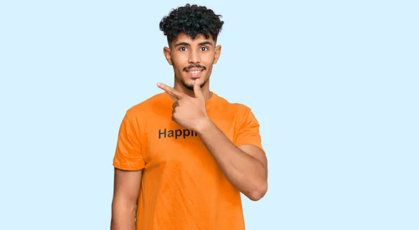 幸せそうな言葉でTシャツを着た若いアラブ人男性が笑顔で陽気に笑顔で手や指を横に向けて幸せと自然な表情で指差す — ストック写真