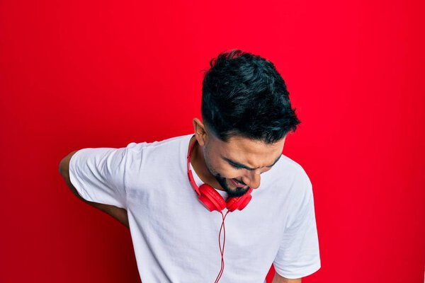 Young man with beard listening to music using headphones suffering of backache, touching back with hand, muscular pain 