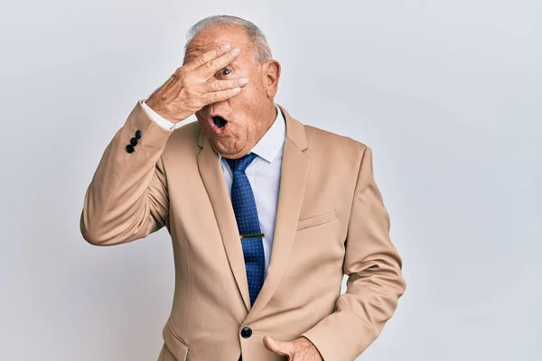 Senior caucasian man wearing business suit peeking in shock covering face and eyes with hand, looking through fingers afraid