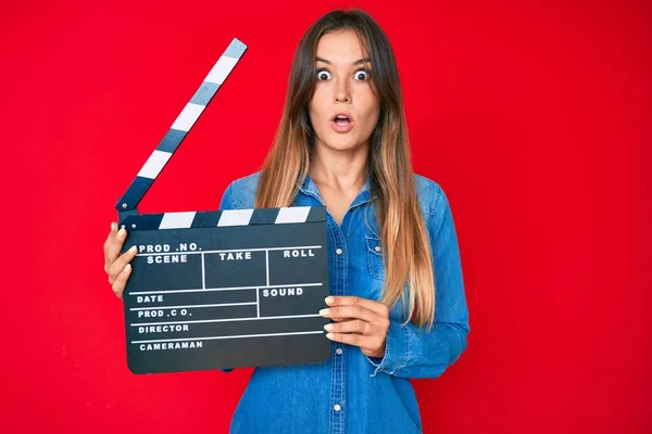 Beautiful caucasian woman holding video film clapboard scared and amazed with open mouth for surprise, disbelief face