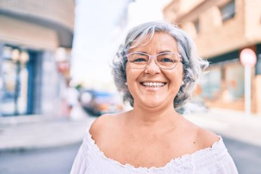 Beyaz saçlı orta yaşlı bir kadın mutlu bir şekilde dışarıda gülümsüyor.