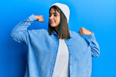 Şirin yün şapka takan genç Latin kadın gururla gülümseyen kol kaslarını gösteriyor. fitness konsepti. 