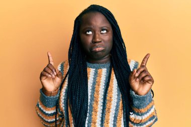 Saçları örgülü genç siyahi bir kadın, sıradan bir kazak giyiyor üzgün ve üzgün görünüyor, parmaklarıyla yönü gösteriyor, mutsuz ve depresif.. 