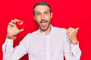 Genç ve yakışıklı bir adam kalbi tutuyor başparmağını yana kaldırıyor mutlu bir şekilde ağzı açık gülümsüyor. 