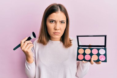 Makyaj fırçası tutan genç ve güzel bir kadın. Yüzü kızardı, şüpheci ve gergin. Problemden dolayı surat asıyor. negatif kişi. 
