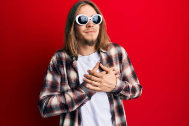 Uzun saçlı, hippi gömleği giyen, güneş gözlüğü takan, göğsünde elleri göğsünde gülümseyen, yüzünde minnet ifadesi olan yakışıklı beyaz bir adam. sağlık kavramı. 