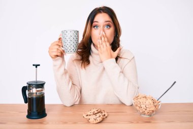 Genç, beyaz, güzel bir kadın masada oturmuş kahvaltısını yapıyor. Ağzını eliyle kapatıyor. Şok olmuş ve hata yapmaktan korkuyor. şaşırmış ifade 