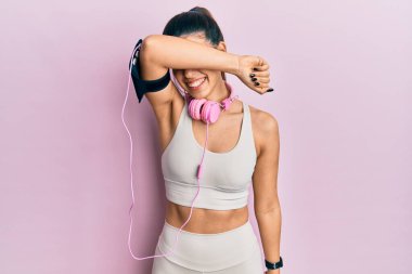 Genç İspanyol kadın spor kıyafetleri giyiyor ve kulaklık takıyor gözleri kollarıyla kapatıyor neşeli ve komik gülümsüyor. kör kavram. 
