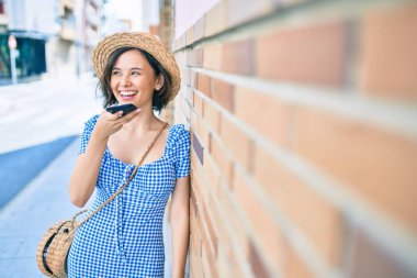 Genç ve güzel bir kız mutlu bir şekilde gülümsüyor. Akıllı telefon kullanıyor ve şehrin duvarına yaslanıyor.