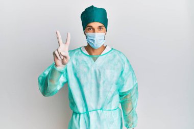 Cerrah üniforması giyen ve tıbbi maske takan genç yakışıklı adam iki numarayı gösterip işaret ederken kendinden emin ve mutlu gülümsüyor.. 