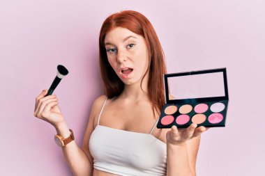 Genç kızıl saçlı kız elinde makyaj fırçası ve yüzü kızaran palet ile kuşkucu ve alaycı görünüyor, ağzı açık kalınca şaşırıyor. 