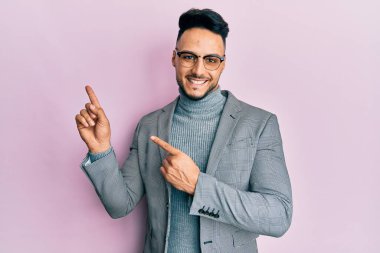 Genç Arap adam iş kıyafetleri içinde gülümsüyor ve kameraya bakıyor iki eliyle ve parmaklarıyla kenarı işaret ediyor.. 